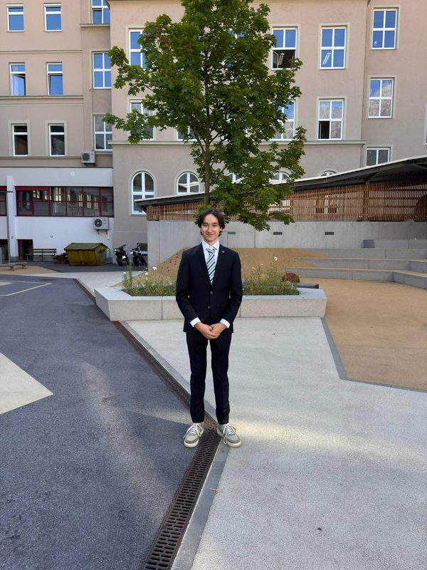 Sandro Exenberger steht in Anzug und gestreifter Krawatte lächelnd in einem gepflasterten Innenhof vor einem Baum und einem mehrstöckigen Gebäude